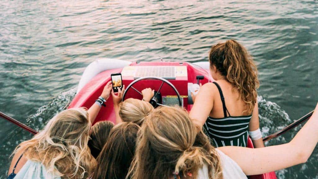 A group of teenage girls are packed into a small boat, while one of them reaches out an arm to take a group selfie.